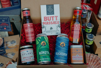 A gift basket containing various bottled products including BBQ sauce, lager, IPA, chilli oil, chicken seasoning, and Gochujang sauce, with branding and labels visible.
