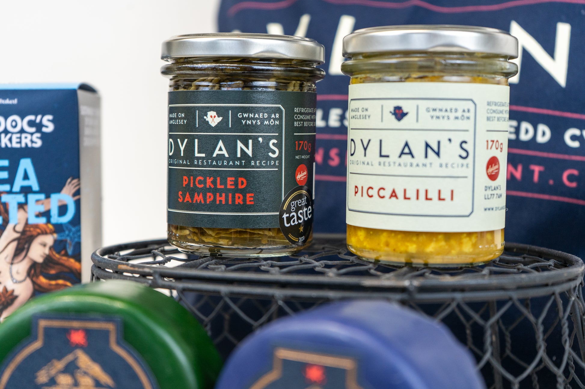 Two jars of Dylan's Pickled Samphire and Piccalilli on a metal stand with branded boxes in the background.