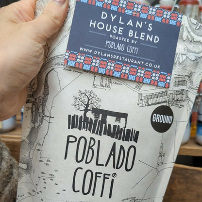 A hand is holding a bag of Dylan's House Blend ground coffee, with a map-like pattern and the brand name 'Poblado Coffi' visible on the packaging.